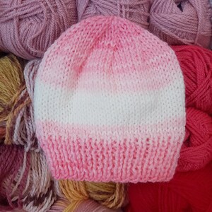 May include: A hand-knitted beanie hat in shades of pink and white. The hat has a ribbed brim and a gradient effect, transitioning from pink to white. The background includes various skeins of yarn in complementary colours.