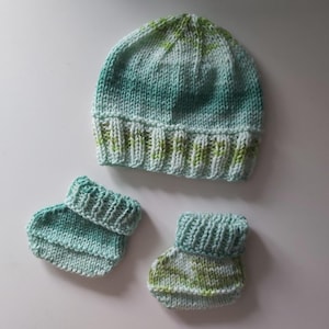 May include: A knitted baby set in shades of teal and green. The set includes a beanie hat and two booties. The hat and booties are made of soft yarn and have a ribbed cuff. The set is designed for a newborn.
