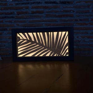 May include: A black framed light fixture with a cutout design of a palm leaf. The light is on and illuminates the leaf design.