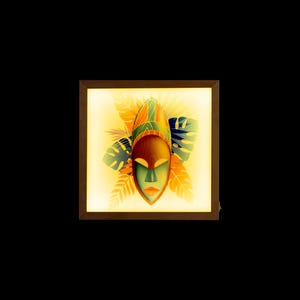 Puede incluir: Una obra de arte enmarcada que presenta una máscara africana colorida con acentos verdes, naranjas y amarillos. La máscara está rodeada de hojas tropicales en tonos verdes y amarillos.