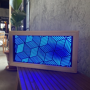 May include: A rectangular wooden framed art piece with a geometric blue light pattern. The pattern is made of intersecting lines that create a cube-like design. The artwork is displayed on a wooden surface.