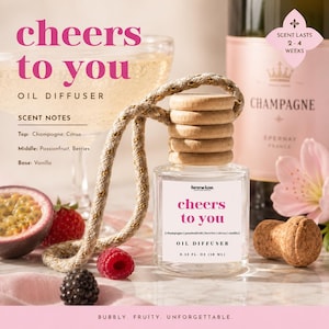May include: Clear glass oil diffuser with a wooden top and a rope hanger. The label reads "cheers to you" with scent notes of champagne, citrus, passionfruit, berries, and vanilla. A bottle of champagne is in the background.