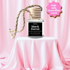 May include: A clear glass oil diffuser bottle with a wooden cap and a woven rope. The label reads "black forest" and "oil diffuser". The bottle sits on a draped pink surface. A pink starburst graphic reads "bold, rich, masculine scent".