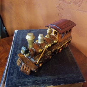 May include: A wooden toy train engine with gold accents and a red roof. The train is sitting on top of a large, dark blue book.