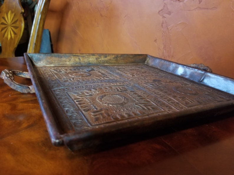 Pode incluir: Uma bandeja de servir de metal vintage com desenhos em relevo ornamentados e duas al&ccedil;as. A bandeja &eacute; marrom escura com uma p&aacute;tina desgastada.