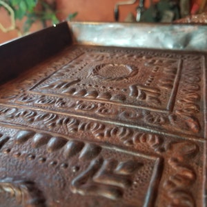 Pode incluir: Um close-up de uma bandeja de metal decorativa com um design em relevo intrincado. A bandeja &eacute; feita de cobre e tem um aspecto r&uacute;stico e envelhecido.