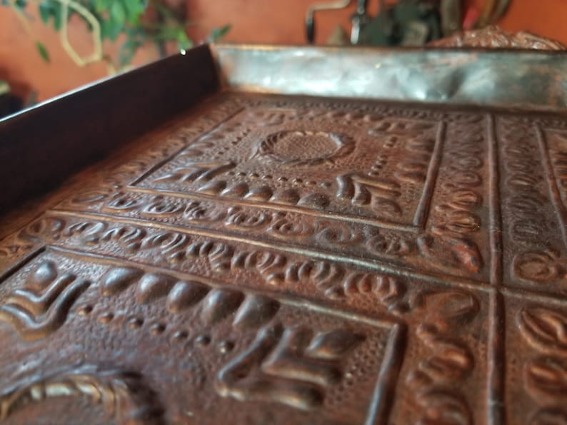 Pode incluir: Um close-up de uma bandeja de metal decorativa com um design em relevo intrincado. A bandeja &eacute; feita de cobre e tem um aspecto r&uacute;stico e envelhecido.
