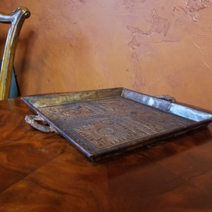Pode incluir: Uma bandeja quadrada de cobre com desenhos em relevo ornamentados e duas al&ccedil;as de metal. A bandeja est&aacute; sobre uma superf&iacute;cie de madeira.