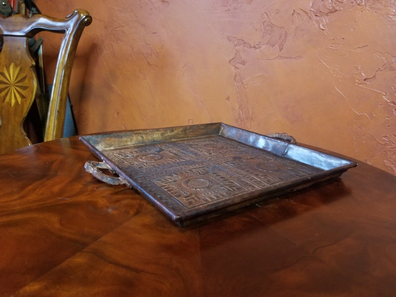 Pode incluir: Uma bandeja quadrada de cobre com desenhos em relevo ornamentados e duas al&ccedil;as de metal. A bandeja est&aacute; sobre uma superf&iacute;cie de madeira.