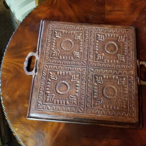 Pode incluir: Uma bandeja quadrada de couro marrom com quatro quadrados decorativos em relevo. A bandeja tem duas al&ccedil;as de metal nas laterais.