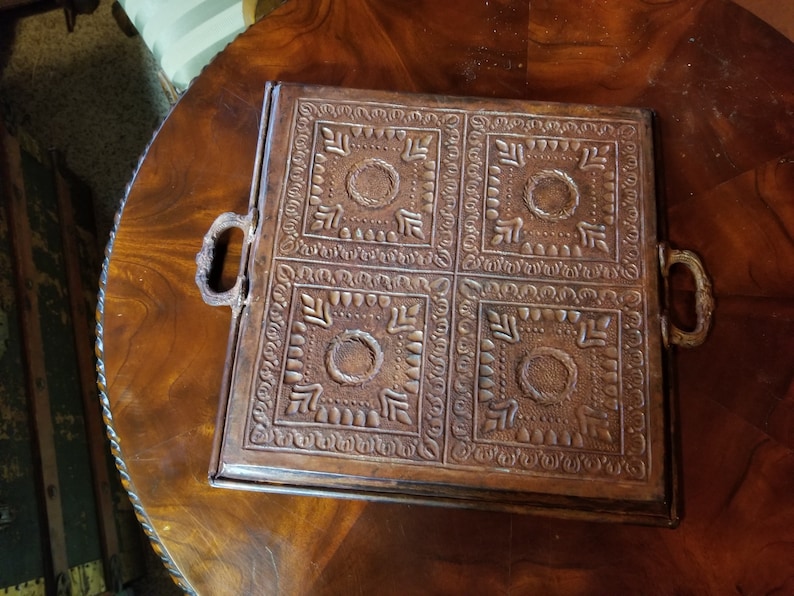 Pode incluir: Uma bandeja quadrada de couro marrom com quatro quadrados decorativos em relevo. A bandeja tem duas al&ccedil;as de metal nas laterais.