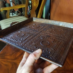 Pode incluir: Uma bandeja de servir quadrada de cobre com desenhos em relevo ornamentados e duas al&ccedil;as. A bandeja &eacute; marrom e tem um aspecto ligeiramente envelhecido.