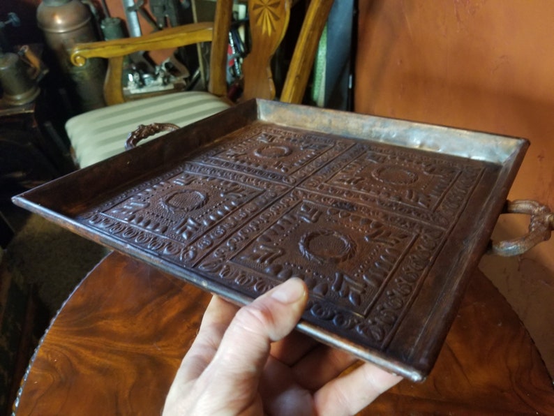 Pode incluir: Uma bandeja de servir quadrada de cobre com desenhos em relevo ornamentados e duas al&ccedil;as. A bandeja &eacute; marrom e tem um aspecto ligeiramente envelhecido.