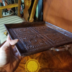 Pode incluir: Uma bandeja quadrada de metal marrom com um design decorativo em relevo. A bandeja tem duas al&ccedil;as de metal nas laterais.