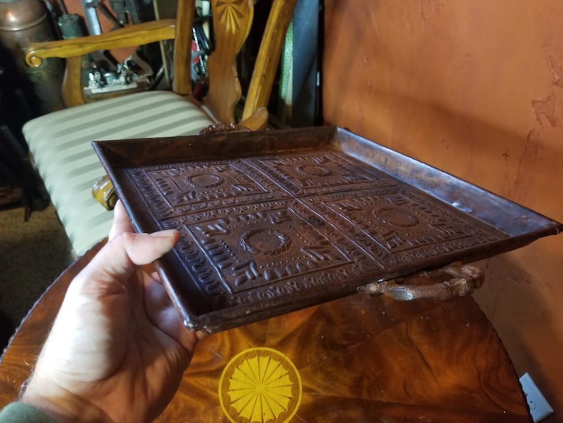 Pode incluir: Uma bandeja quadrada de metal marrom com um design decorativo em relevo. A bandeja tem duas al&ccedil;as de metal nas laterais.