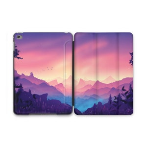 Puede incluir: Una puesta de sol púrpura y rosa sobre una cordillera con una silueta de árboles y un pequeño edificio. La imagen está impresa en una funda negra para iPad.
