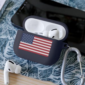 May include: A navy blue silicone case for wireless earbuds with a printed American flag design. The case has a carabiner clip attached.