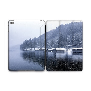 May include: A snowy winter scene with a lake and a row of small cabins along the shore. The trees in the background are covered in snow and the lake is calm and still.