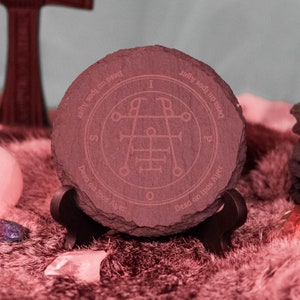 May include: A dark gray slate coaster with a circular design featuring a sigil, the text "Desa an Ipos Ager", and the letters "I.S.P."