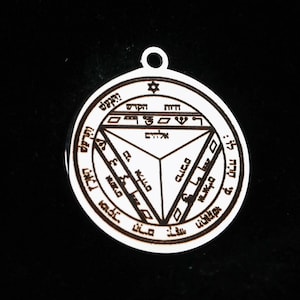 Può includere: Un ciondolo circolare in metallo color argento con un design geometrico inciso che presenta un triangolo centrale, una piccola stella in alto e vari caratteri ebraici inscritti all'interno del bordo circolare e delle sezioni triangolari.
