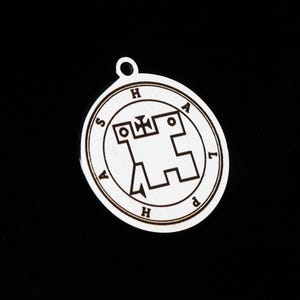 May include: A circular silver-toned metal pendant featuring a central engraved geometric sigil design. The outer border of the pendant contains the engraved letters S, H, A, L, P, H, A, H arranged in a circle. A small loop is attached at the top.