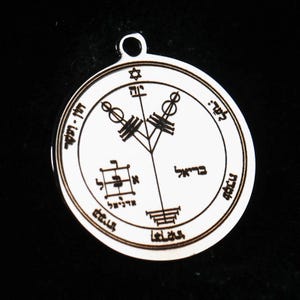 May include: A circular silver-toned metal pendant featuring an engraved design with a central vertical line, two branching symbols, a small Star of David at the top, a square grid, and Hebrew text inscribed along the inner and outer circular borders.