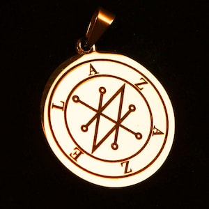 May include: A circular gold-toned metal pendant featuring a central engraved sigil with radiating lines ending in small circles. The perimeter of the pendant is inscribed with the letters A, Z, A, Z, E, L in a circular arrangement around the sigil.
