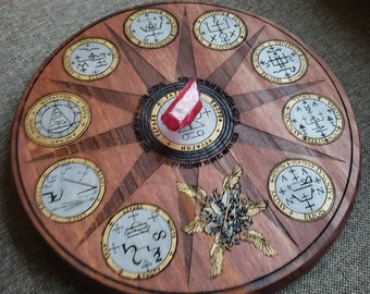 May include: A wooden circular board with a compass design featuring eight sections. Each section has a different symbol and text in black and white. The center of the board has a black and white symbol with the text "ARIATON", "KROPER", and "FAZL" in black. A red piece of paper is placed on the board.