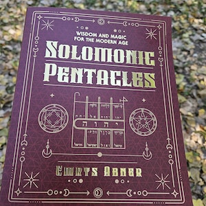 Puede incluir: Un libro granate con letras e ilustraciones doradas. El t&iacute;tulo es "Solomonic Pentacles", con el subt&iacute;tulo "Wisdom and Magic for the Modern Age". Tambi&eacute;n se ve el nombre del autor, Emrys Abner. El libro se sostiene al aire libre.