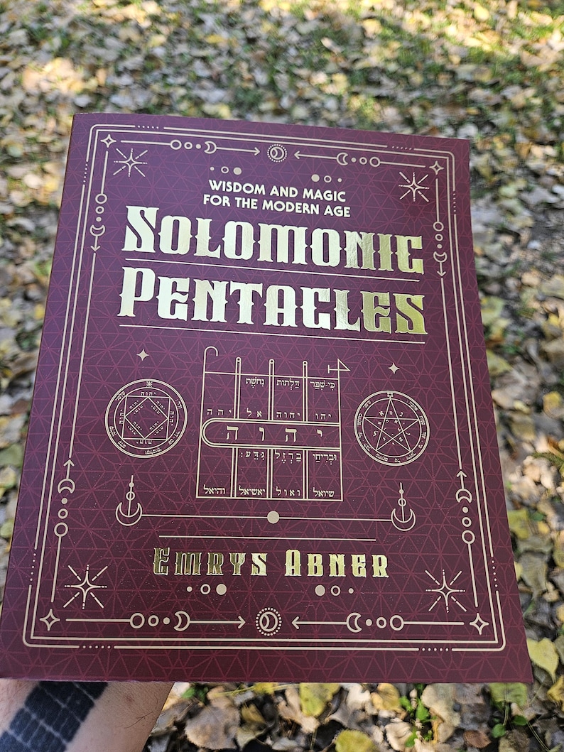 Puede incluir: Un libro granate con letras e ilustraciones doradas. El t&iacute;tulo es "Solomonic Pentacles", con el subt&iacute;tulo "Wisdom and Magic for the Modern Age". Tambi&eacute;n se ve el nombre del autor, Emrys Abner. El libro se sostiene al aire libre.