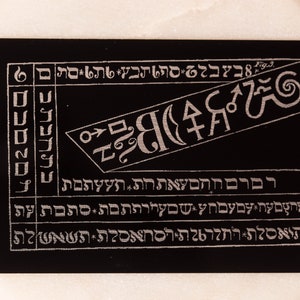 Puede incluir: Una tarjeta negra con texto y símbolos plateados. El texto incluye letras y números hebreos. La tarjeta tiene un diseño de espiral y un símbolo que parece una luna creciente con una estrella en su interior.