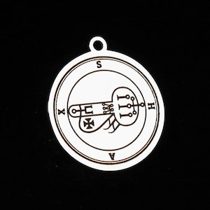 Pode incluir: Um pingente circular branco com um selo gravado escuro no centro. O design inclui um emblema central com linhas e uma cruz, rodeado por uma borda de linha dupla. As letras S, H, A e X estão posicionadas ao redor do perímetro.