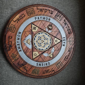 May include: A circular wooden plaque with a silver circle in the center. The silver circle features a triangle with an eye inside, surrounded by other symbols. The plaque is inscribed with the words "PAYMON", "AMAYMON", "EGYN", "ORIENS", and other text in a foreign language.