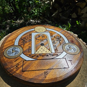 May include: A wooden round disc with a silver and gold design. The design features a circle with a triangle inside, a square with a circle inside, and a rectangle with a circle inside. The disc also has a blank rectangle in the center. The disc is surrounded by a circle of text in a foreign language.