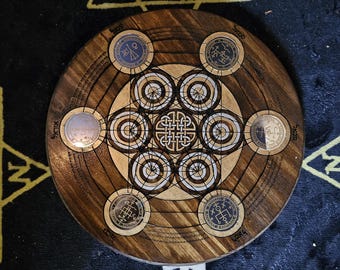 May include: A wooden circular board with a geometric design in the center. The board has six circular metal pieces with symbols and text around the edge. The board is brown with a light wood grain.