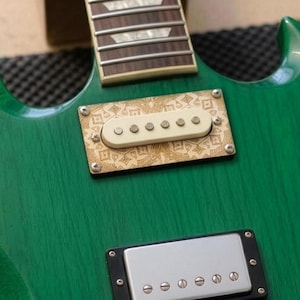 Könnte beinhalten: Nahaufnahme einer grünen E-Gitarre. Die Gitarre hat einen Holz-Hals mit silbernen Bünden und einen grünen Korpus. Die Gitarre hat zwei Tonabnehmer, einen mit cremefarbenem Deckel und einen mit silbernem Deckel. Die Brücke ist silberfarben.