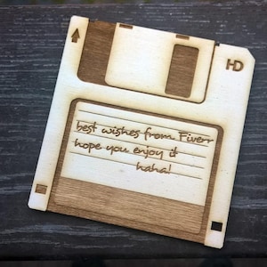 Op de afbeelding: Een houten voorwerp in de vorm van een diskette met de tekst "best wishes from Fiverr hope you enjoy it haha!". Het item is gemaakt van licht en donkerbruin hout, met de tekst gegraveerd.