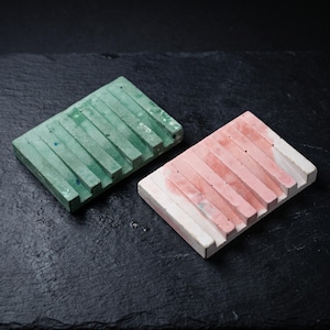 Puede incluir: Dos jaboneras rectangulares de hormigón, una en un tono verde apagado y la otra en un tono rosa suave. Cada jabonera presenta una serie de crestas paralelas elevadas para el drenaje, con un acabado de superficie texturizado y poroso.