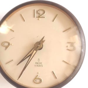 May include: A vintage round clock with a cream-colored face and gold-colored hands and numerals. The clock has a dark brown frame and the text "8 ANCRE 11 RUBIS" is visible near the center. The clock face shows the numbers 12, 3, 6, and 9.