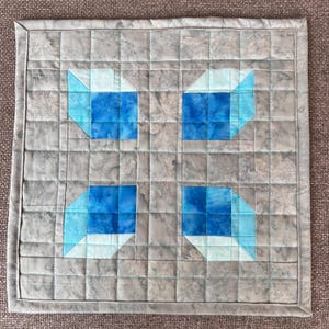 May include: A gray quilted fabric square with a blue and white geometric pattern. The pattern is made up of four squares with a white center and blue edges.