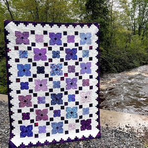 May include: A rectangular quilt featuring a repeating pattern of four-petaled flower shapes in various shades of purple, blue, and violet. The floral blocks are separated by a white grid and bordered by a dark purple and white geometric chain design.