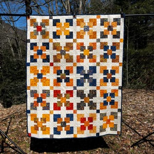 May include: A handmade quilt featuring a repeating pattern of squares in orange, yellow, blue, and red on a white backdrop. The quilt is displayed outdoors, highlighting its detailed design and craftsmanship. A cozy home decor item.