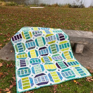 May include: A colorful quilt with a geometric pattern of teal, green, yellow, and purple shapes. The quilt is displayed outdoors on a stone bench and grass, with a lake and trees in the background. The quilt has a white border.