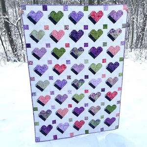 May include: A handmade quilt featuring a repeating pattern of 3D hearts in shades of purple, pink, green, and red on a white background. The quilt has a geometric design with square accents and a purple border. The quilt is held up in front of a snowy background.