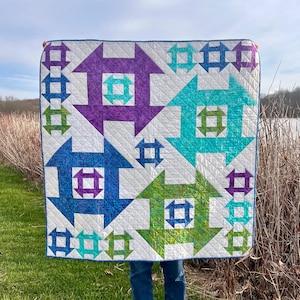 May include: A white quilt with a geometric pattern of blue, green, and purple arrows. The arrows are made up of smaller squares and rectangles.