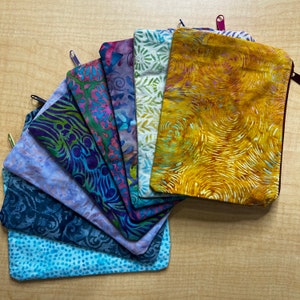 May include: A set of nine colorful zippered pouches made of fabric with various patterns. The pouches are arranged in a fan shape, with the largest pouch at the top and the smallest pouch at the bottom.