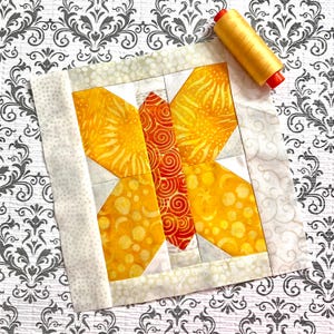 May include: A square fabric quilt block featuring a central butterfly motif constructed from yellow and orange patterned fabric pieces, surrounded by a light cream border with a subtle swirl design, accompanied by a spool of yellow sewing thread.