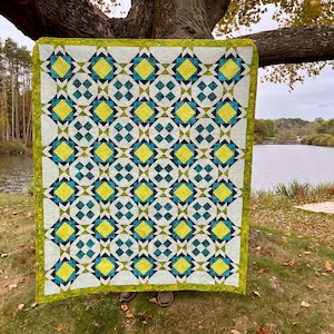 May include: A handmade quilt with a geometric pattern in shades of yellow, teal, and navy blue on a white background. The quilt is bordered with a green fabric and is displayed outdoors, hanging from a tree branch.