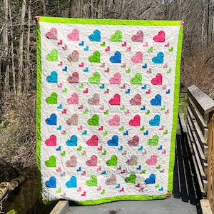 May include: A rectangular quilt featuring a white background with a swirling quilted pattern, decorated with a repeating grid of heart shapes in shades of pink, blue, and green, finished with a bright lime green border around the entire edge.