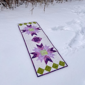 Peut inclure: Un chemin de table matelassé violet et vert avec un motif en étoile. Le chemin de table est posé sur une surface enneigée.